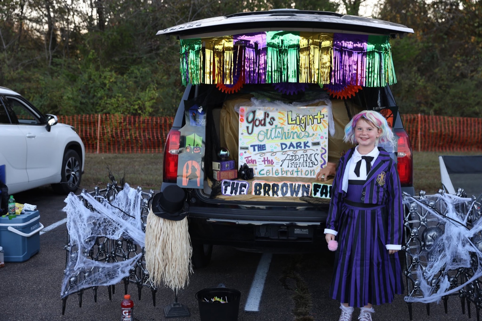 An SUV trunk with purple and green fringe and a sign reading God's Light Outshines the Dark, a girl dressed as Wednesday Addams in front.