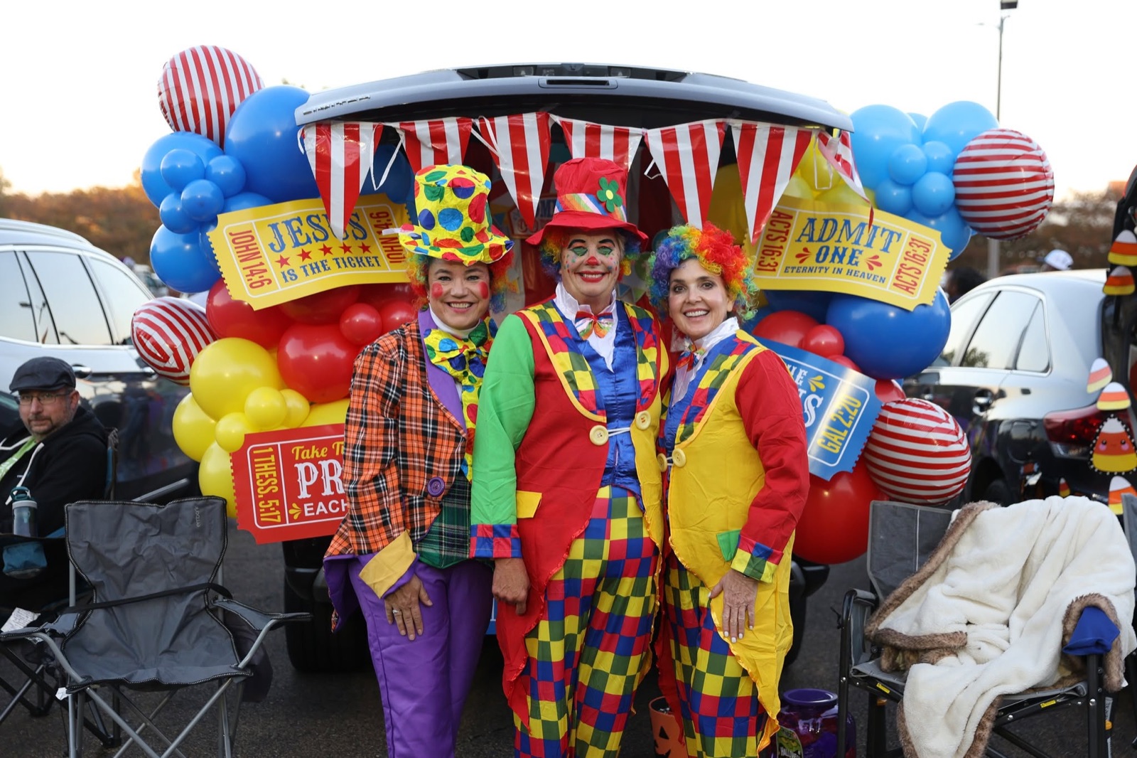 Three people dressed as clowns in front of a trunk decorated like a circus tent with red, yellow and blue balloons.