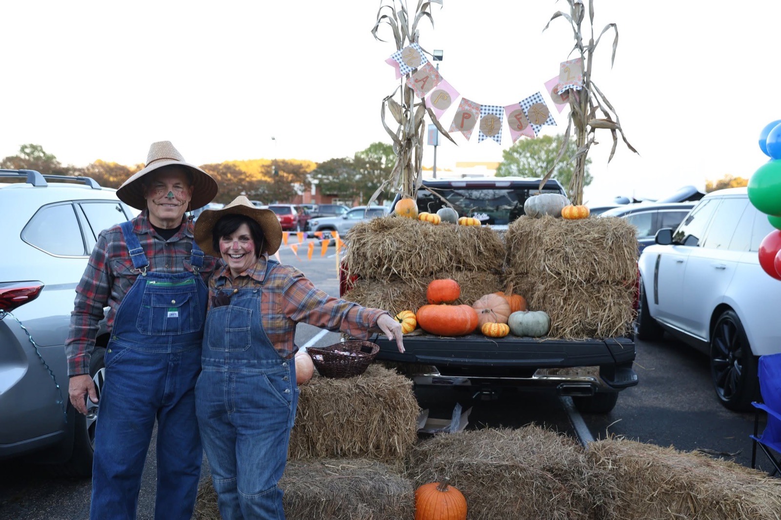 A couple in overalls and straw hats beside a truck bed with hay bales, pumpkins and corn stalks.