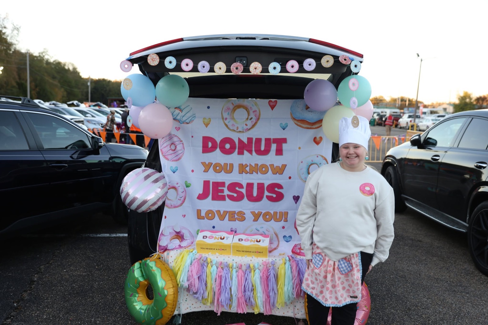 A trunk decorated with pastel donuts and a sign reading Donut You Know Jesus Loves You, a woman in a baker's hat beside it.